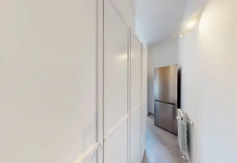 Narrow kitchen or utility corridor with tall white cabinets along one side and a stainless-steel refrigerator at the end. Simple, functional layout suitable for compact apartments.