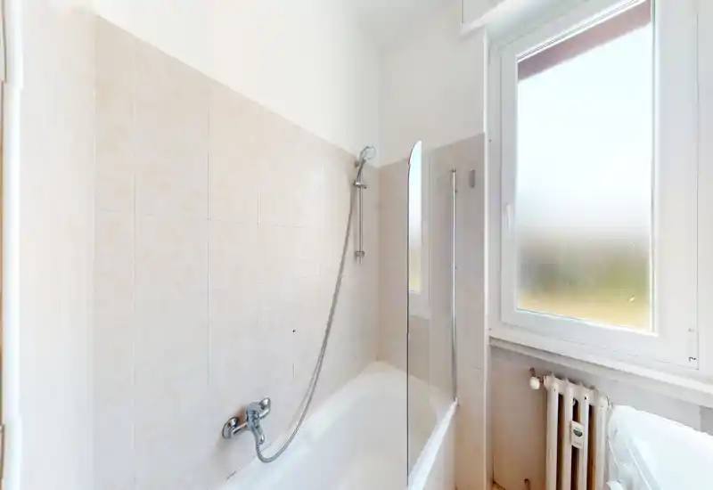 Bright, clean bathroom with a bathtub, handheld shower and partial glass screen next to a frosted window; neutral tile walls and a radiator are visible.