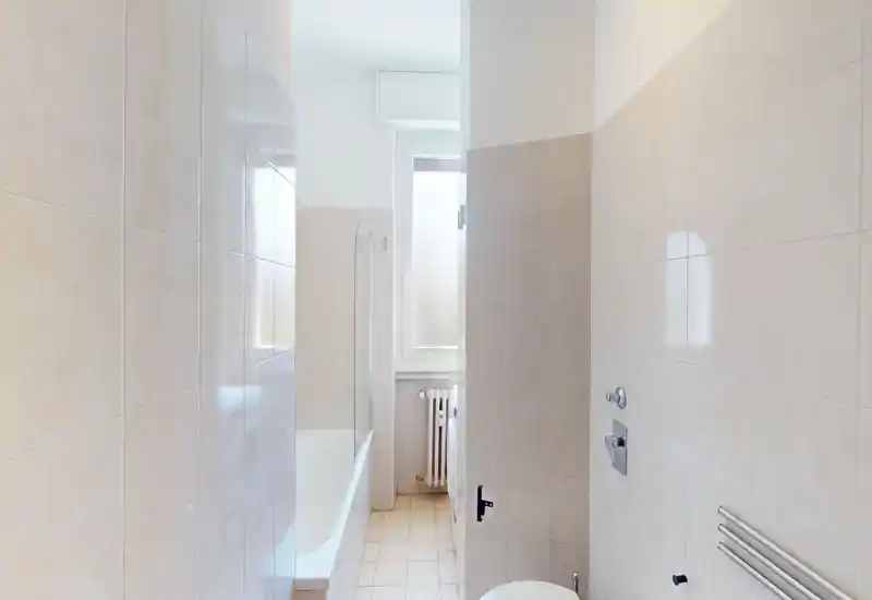 Narrow, tiled bathroom with a bathtub on the left, wall-mounted radiator and a toilet partially visible; bright natural light from a frosted window.