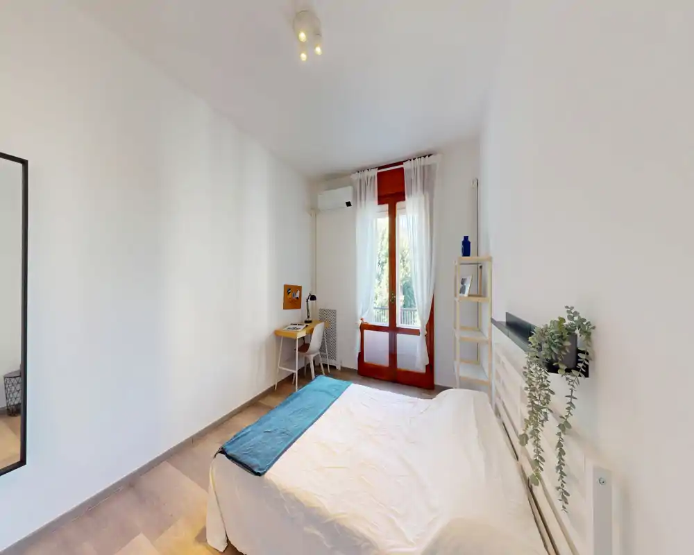 Chambre lumineuse et minimaliste avec un lit double, lumière naturelle par une porte-fenêtre et un petit bureau près de la fenêtre. Décor sobre, étagères et plante suspendue pour une ambiance calme, idéale pour une annonce.