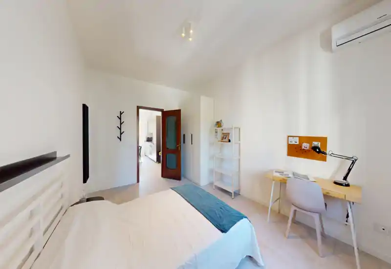 Dormitorio luminoso y minimalista con cama doble, estante a modo de mesita y un pequeño escritorio junto a la pared. Tonos neutros y buena luz natural, ideal para alquileres temporales o estudiantes.