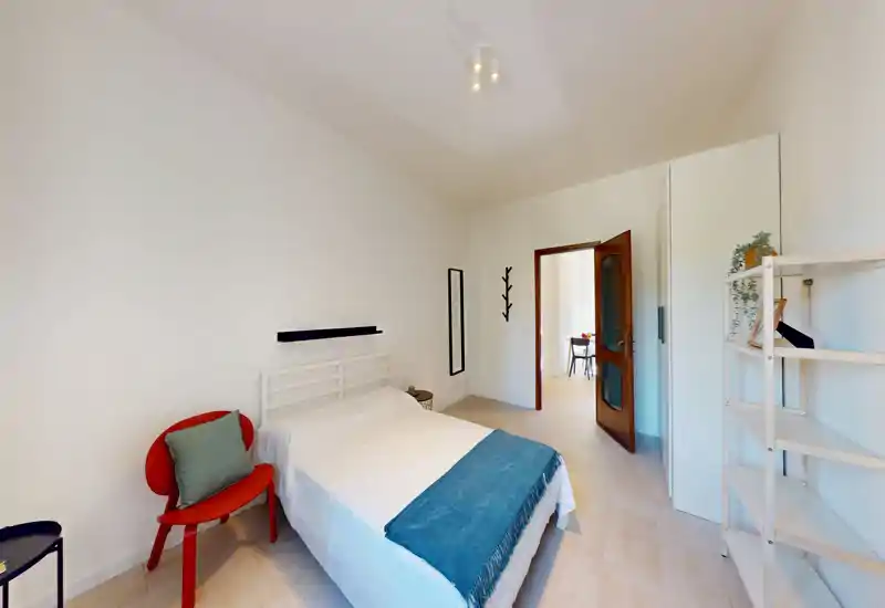 Dormitorio luminoso y minimalista con cama doble bien hecha, suelo claro y una pequeña butaca. Líneas limpias y textiles suaves ofrecen un espacio tranquilo y acogedor para el anuncio.