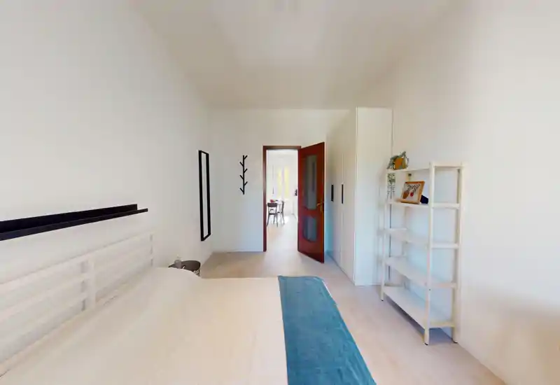 Dormitorio luminoso y minimalista con cama doble visible, estantería y armario sencillos; tonos neutros y una banda azul que aportan una sensación tranquila y aireada.
