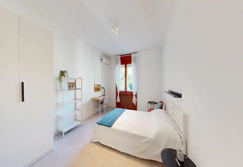 Dormitorio luminoso y minimalista con cama doble, mesitas y un pequeño escritorio junto a la ventana. La luz natural y las paredes blancas crean un ambiente tranquilo y aireado.