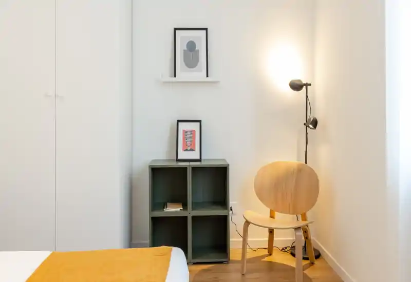 Angolo camera luminoso e minimal con una sedia in legno, scaffalatura accanto al letto e parte del letto con un caldo plaid ocra; pareti neutre e una lampada da terra creano un'atmosfera accogliente e moderna.