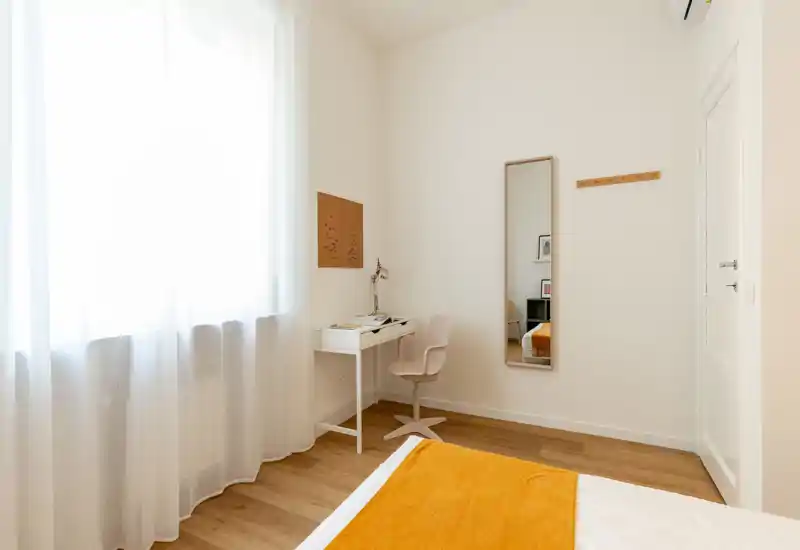 Angolo camera da letto luminoso e minimale con una scrivania piccola, uno specchio a figura intera e il letto parzialmente visibile con un caldo copriletto arancione. La luce naturale attraversa le tende leggere, creando un'atmosfera contemporanea e tranquilla.
