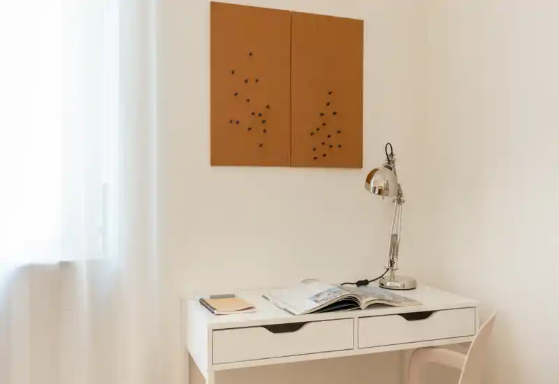 Angolo scrivania luminoso e minimale con tavolo bianco, lampada cromata, pannelli in sughero e luce naturale soffusa — ideale come piccolo home office o area studio.