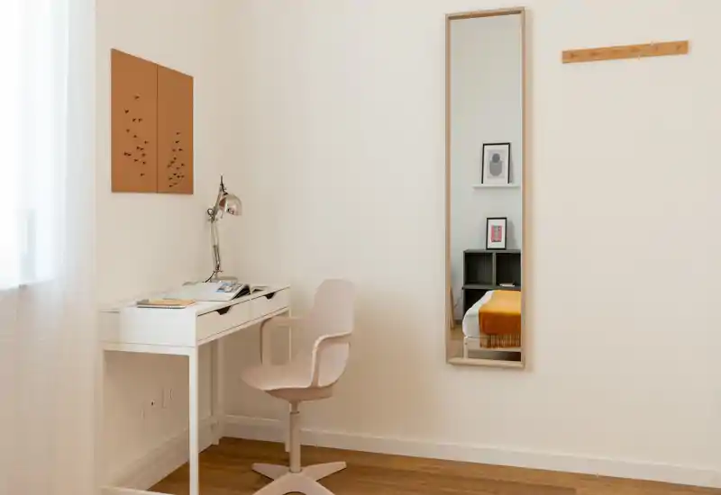 Angolo camera luminoso e minimale con scrivania e sedia; il letto si vede nel riflesso di uno specchio alto, che aggiunge profondità. Luce naturale e toni neutri creano un ambiente tranquillo e funzionale, ideale per una persona.