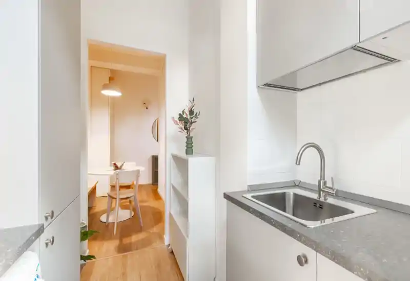 Angolo cucina moderno e compatto con lavello in acciaio, piano grigio e mobili bianchi: ambiente luminoso e ben arredato che si apre su un piccolo angolo pranzo.