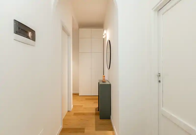 Ingresso-corridoio luminoso e minimale con pavimento in legno, un sottile tavolino consolle e specchio a parete, che conduce a armadi a muro.