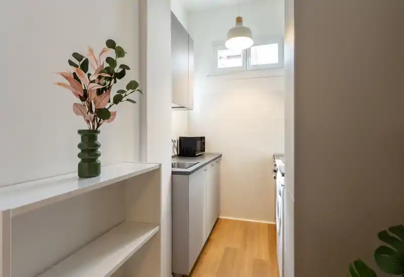 Cucina compatta a corridoio con pensili grigi, lavello, microonde e lavatrice; luminosa, pulita e organizzata con efficienza per spazi piccoli.