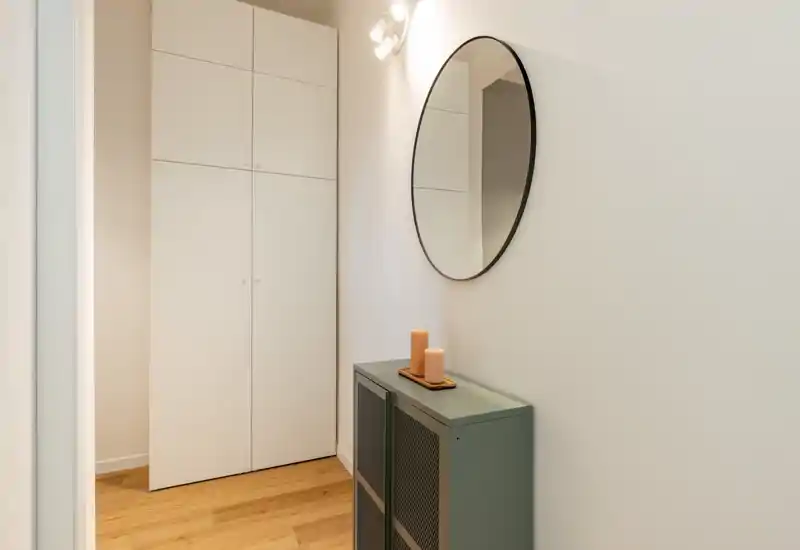 Ingresso ordinato e ben illuminato con un sottile mobile consolle, specchio rotondo a parete e armadi a muro; il pavimento in legno e l'arredo minimal creano una prima impressione accogliente.