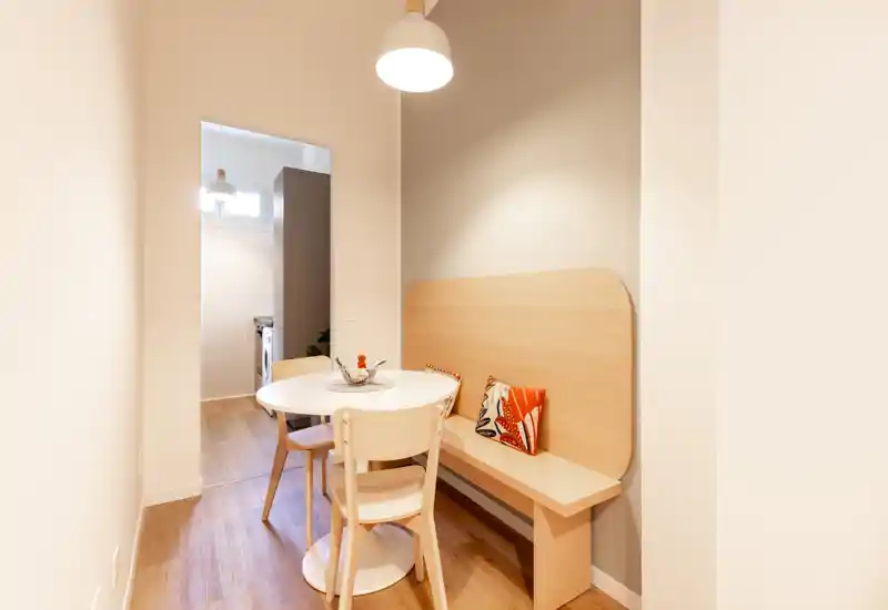 Angolo pranzo compatto con tavolo rotondo, panca e sedie in legno, illuminato da un lampadario a sospensione; pavimenti in legno e arredo essenziale creano un ambiente accogliente.