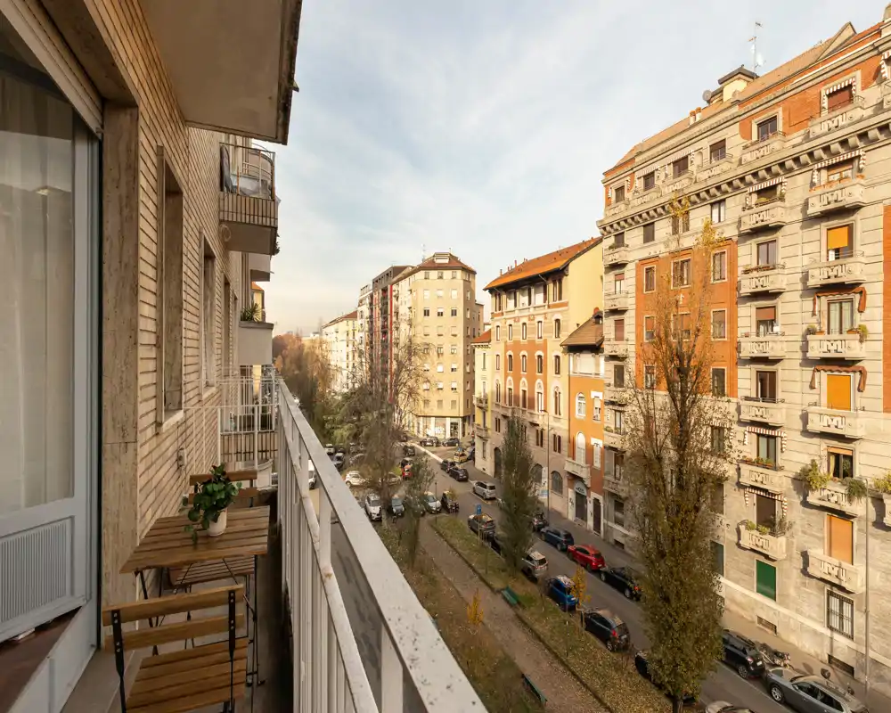 Balcone soleggiato con vista su una strada alberata e palazzi storici, dotato di un piccolo tavolo e sedie in legno — perfetto per il caffè mattutino.