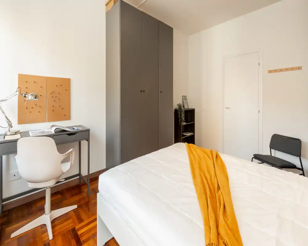 Camera da letto luminosa e ordinata con un ampio letto in primo piano, una scrivania sottile con sedia e un armadio alto contro la parete. Il pavimento in legno caldo e il plaid giallo donano un tocco accogliente all'arredamento minimal.