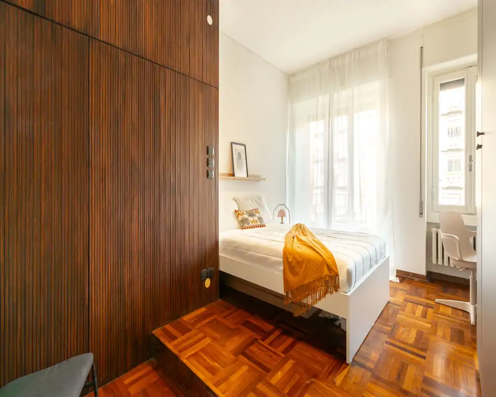 Camera da letto luminosa e ben arredata con letto matrimoniale, parete effetto legno e pavimento in parquet. Ampia finestra che illumina l'ambiente; plaid e cuscini decorativi rendono lo spazio accogliente.