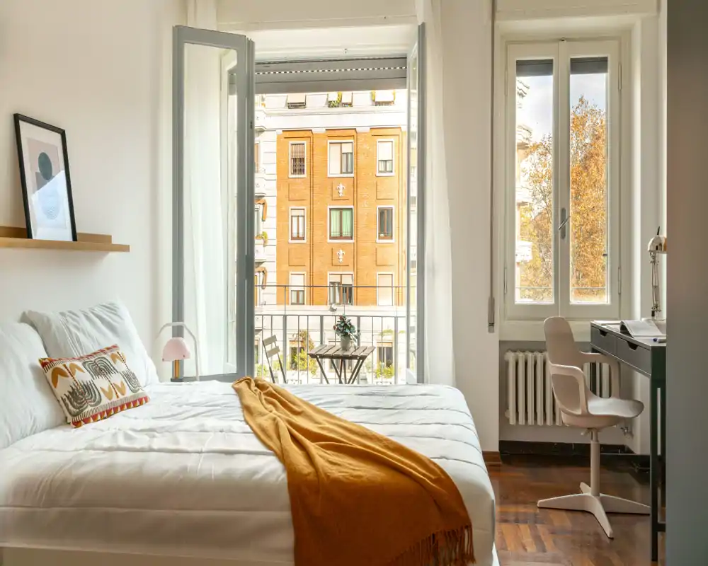 Camera da letto luminosa e di stile con un grande letto con biancheria bianca, plaid ocra e cuscini decorativi; una piccola scrivania vicino alla finestra e un balcone con tavolino che offre una gradevole vista sulla città.