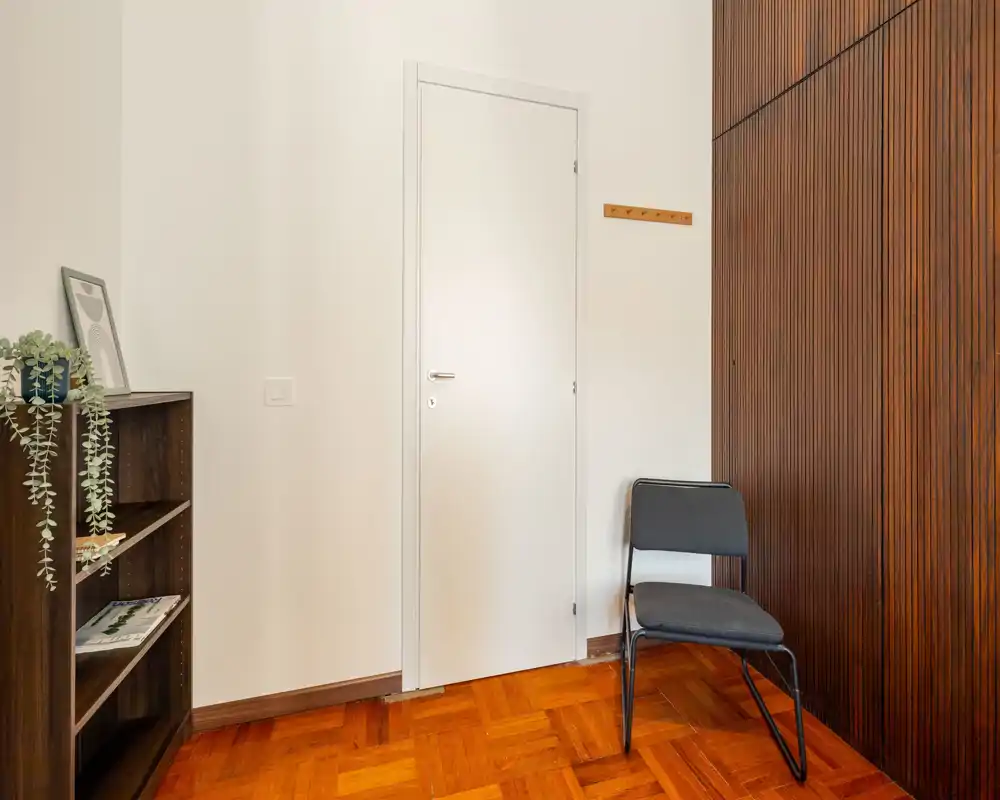 Piccolo ingresso ordinato con porta bianca chiusa, una sedia semplice e una libreria in legno; il pavimento in parquet caldo e le doghe verticali in legno danno carattere.