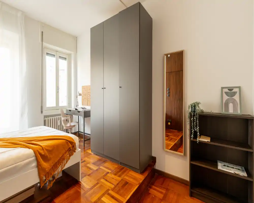 Camera da letto luminosa e ben arredata con letto singolo comodo, armadio e una piccola scrivania vicino alla finestra. Il parquet caldo e i tessili creano un’atmosfera accogliente, perfetta per gli annunci.