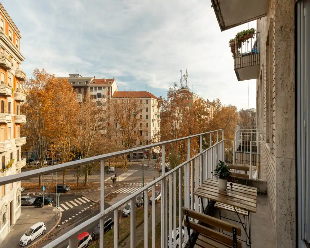 Balcone urbano soleggiato con tavolino e sedie in legno, pianta in vaso e una vista ampia sulla strada incorniciata da alberi autunnali — ideale per il caffè mattutino.