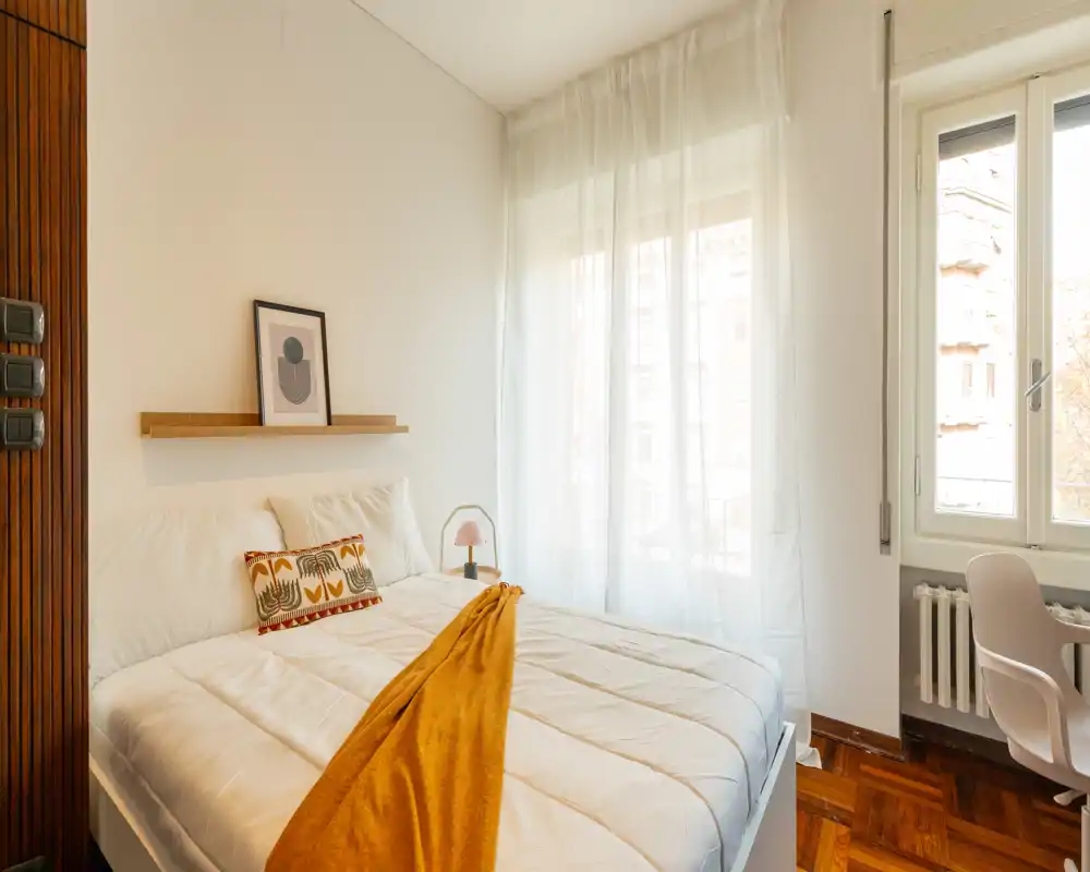 Camera da letto luminosa e accogliente con letto grande con biancheria bianca, plaid senape, cuscino decorativo e tende leggere che diffondono la luce naturale.