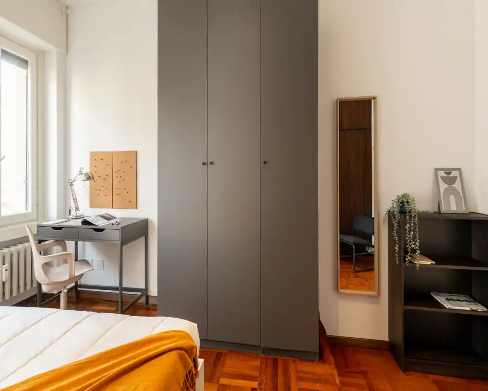 Camera da letto luminosa e moderna con letto parzialmente visibile, armadio a muro, scrivania vicino alla finestra e pavimento in legno caldo — arredata con un accogliente plaid ocra.