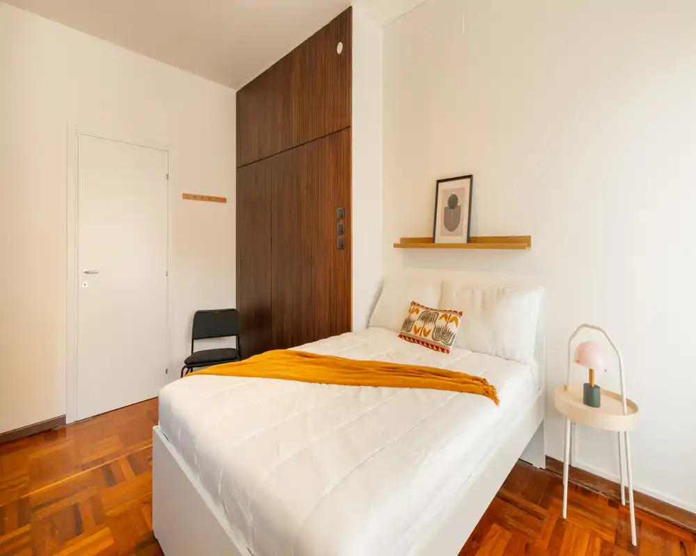 Camera da letto luminosa e minimalista con letto ben rifatto, pavimento in legno caldo e un piccolo comodino con lampada — perfetta per un soggiorno accogliente.