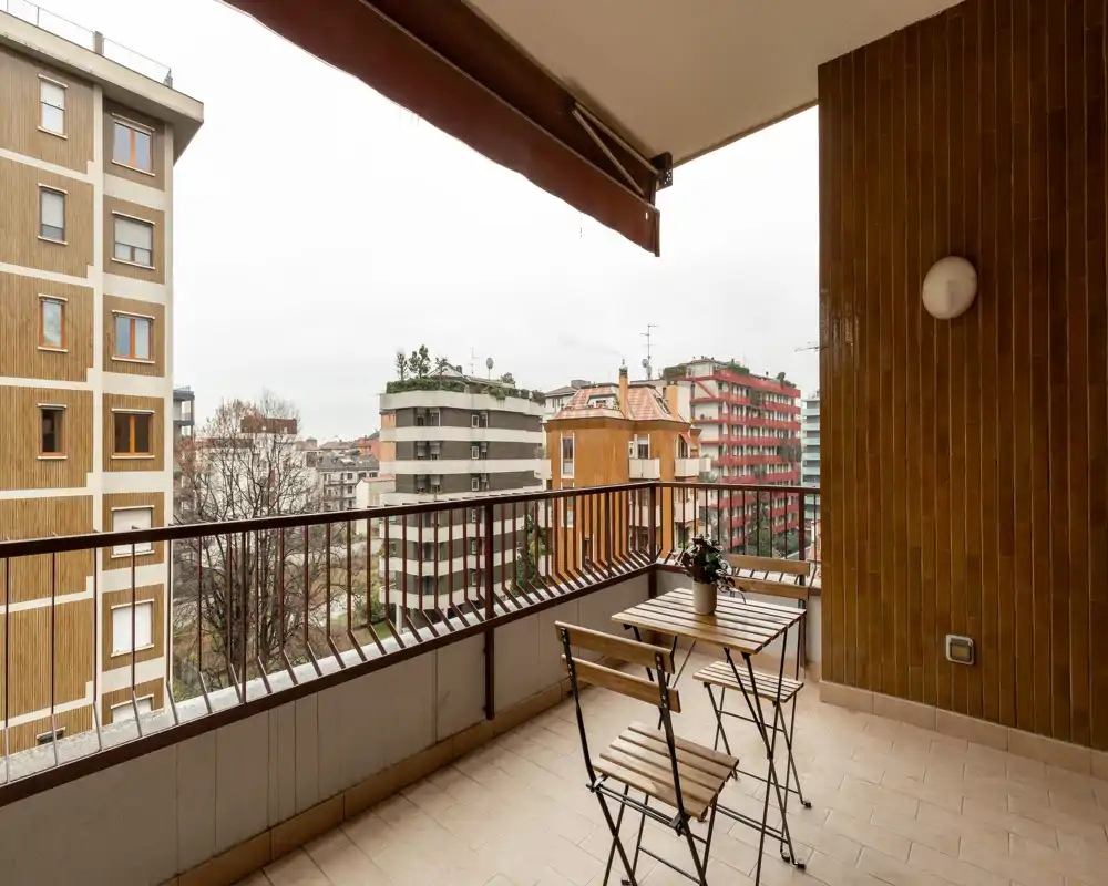 Balcón cubierto con pequeña mesa bistró de madera y dos sillas plegables, suelo de baldosa y vistas a la ciudad — ideal para el desayuno.
