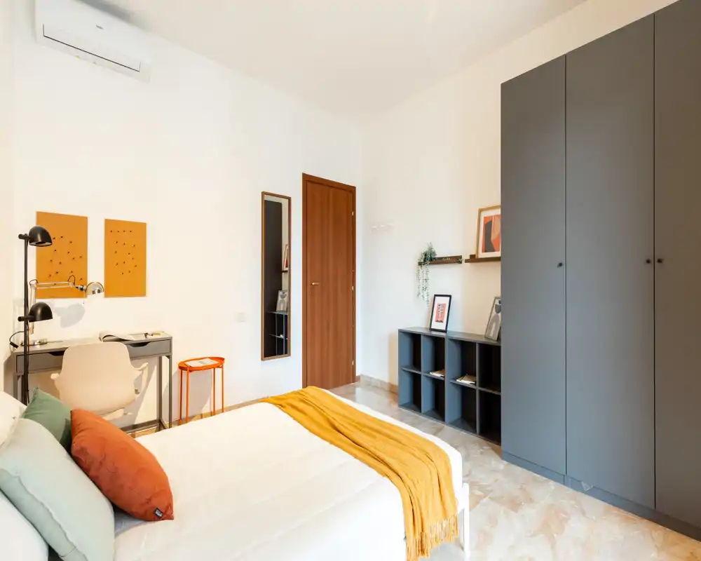 Dormitorio luminoso y bien decorado con cama doble cómoda, cojines de colores y una manta mostaza. La habitación cuenta con escritorio, armario y estanterías, ideal para descansar y trabajar de forma ligera.