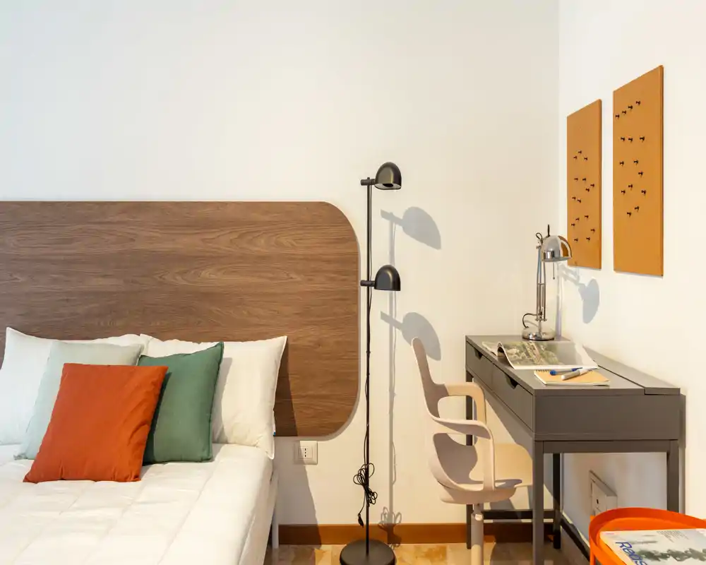 Dormitorio moderno y luminoso con cabecero de madera, ropa de cama blanca y cojines de color; a la derecha hay un escritorio con lámpara que crea un rincón funcional para dormir y trabajar.