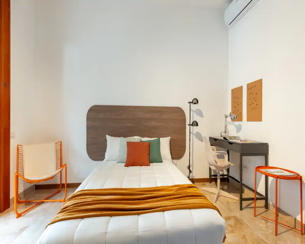 Dormitorio moderno y luminoso con cama completa, manta cálida y un espacio de trabajo compacto con escritorio y silla — ideal para una estancia cómoda.