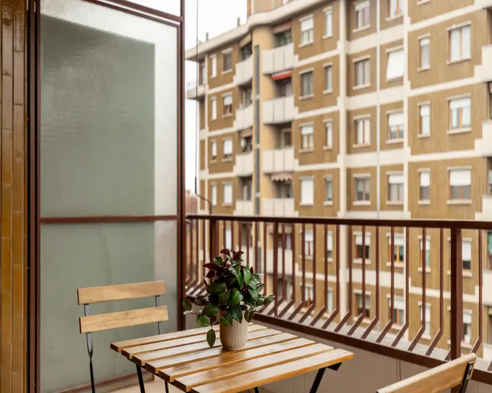 Balcón compacto y soleado con mesa de madera y sillas plegables, planta en maceta sobre la mesa y vistas a la ciudad — ideal para el café de la mañana.