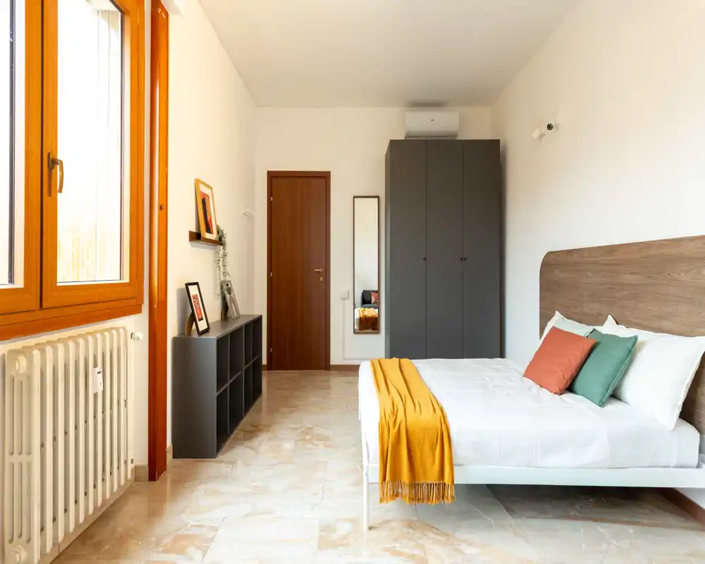 Dormitorio moderno y luminoso con cabecero de madera, cama doble bien hecha con cojines de colores y manta mostaza, ventana grande que aporta luz natural, armario y estantería simple para almacenamiento.