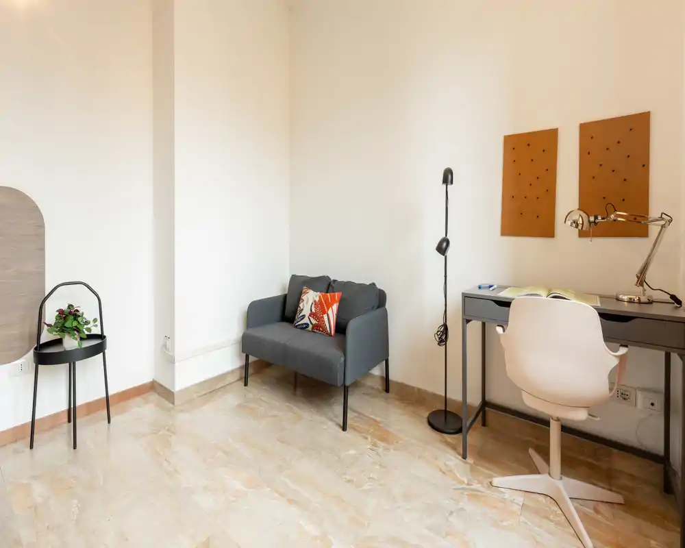 Área de estar minimalista y bien iluminada con un pequeño sofá de dos plazas, un escritorio y una silla, y detalles decorativos sencillos — ideal para un estudio compacto o rincón de trabajo.