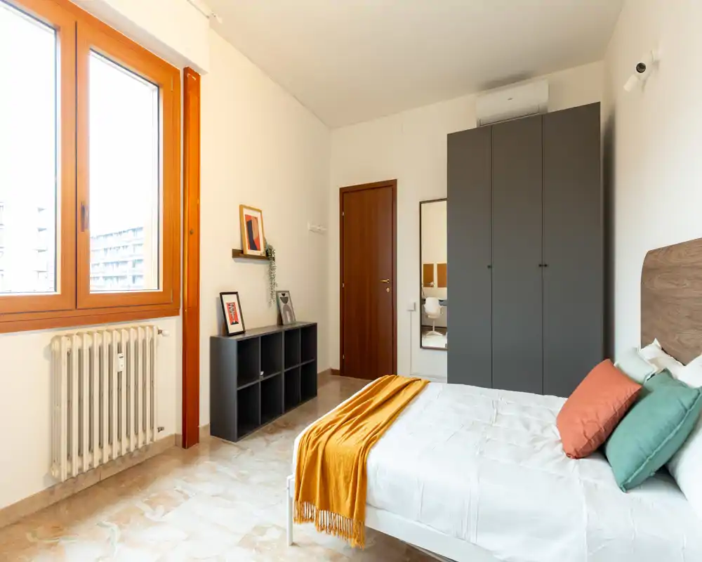Dormitorio moderno y luminoso con cama bien hecha, cojines de colores y una manta mostaza, un armario grande y una ventana que aporta mucha luz natural.