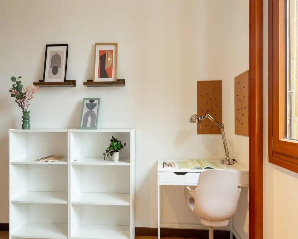 Angolo luminoso e minimale con scaffali bianchi, una piccola scrivania e stampe decorative. La luce naturale dalla finestra rende lo spazio arioso e adatto allo studio o al relax.