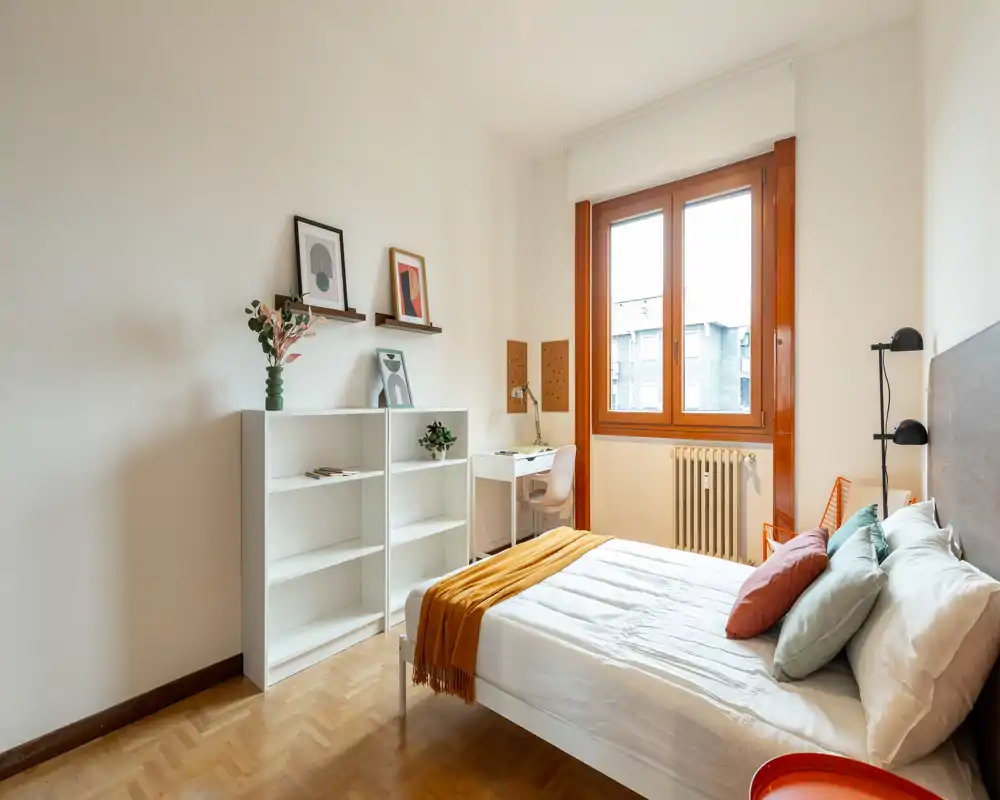 Camera da letto luminosa e ben arredata con letto matrimoniale, cuscini e plaid senape. Una scrivania vicino alla finestra e scaffalature aperte rendono lo spazio pratico e accogliente, ideale per affitti brevi.