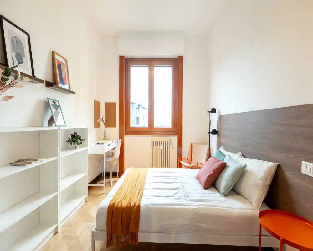 Camera da letto luminosa e ben arredata con letto matrimoniale, testiera in legno, cuscini colorati e plaid color senape. Una scrivania vicino alla finestra e scaffalature offrono spazio di lavoro e storage, perfetta per annunci in affitto.