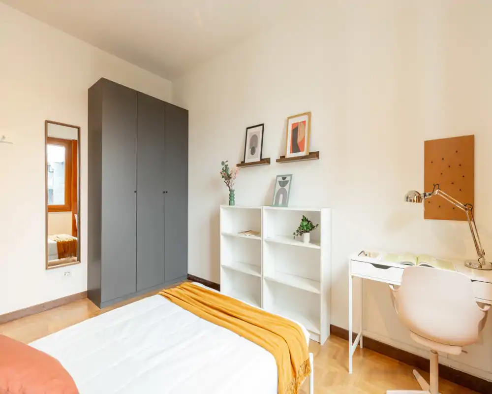 Camera da letto moderna e luminosa con letto ben rifatto, plaid color senape, armadio scuro, mensole e una piccola scrivania — ideale per una persona o uno studente.