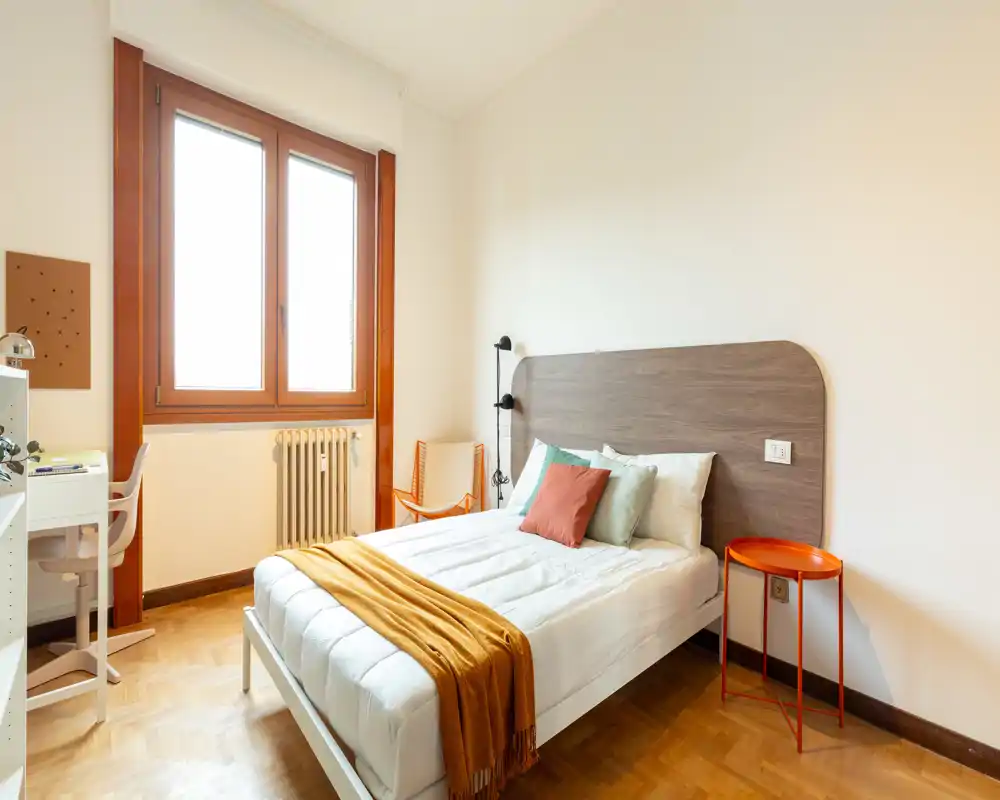 Camera da letto luminosa e curata con un letto matrimoniale, testiera in legno e luce naturale da una grande finestra. Angolo scrivania e comodino rendono la stanza funzionale per riposo e studio.