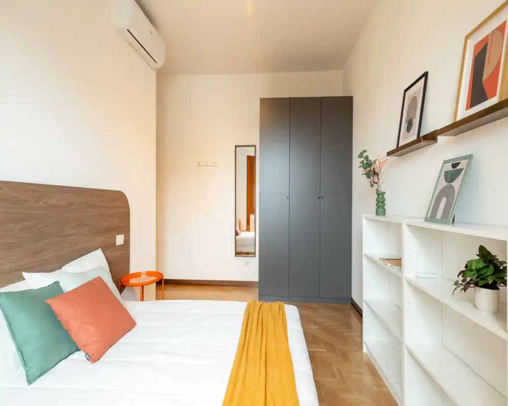 Camera da letto moderna e luminosa con un ampio letto, cuscini colorati e un caldo plaid. L'ambiente include un armadio, scaffali con quadri decorativi e un piccolo comodino: accogliente e ben illuminata, perfetta per annunci.