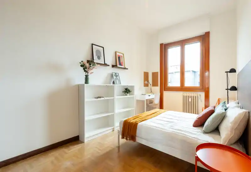 Dormitorio luminoso y bien decorado con cama doble, cojines de colores y una manta mostaza; ventana grande que aporta mucha luz natural y una pequeña mesa de estudio junto al radiador.