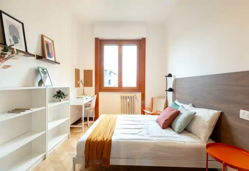 Dormitorio luminoso y bien decorado con cama doble, cabecero de madera, cojines de colores y manta mostaza. Un escritorio junto a la ventana y estanterías ofrecen espacio de trabajo y almacenamiento, ideal para anuncios de alquiler.