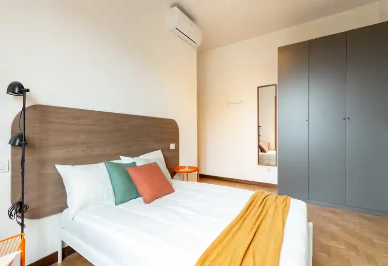 Dormitorio moderno y luminoso con cabecero de madera, ropa de cama blanca, cojines coloridos y una manta mostaza, armario empotrado y espejo de cuerpo entero.