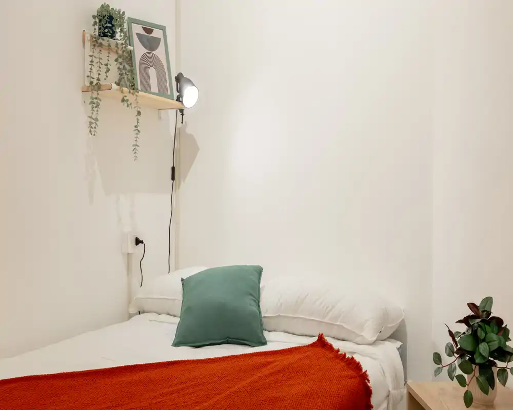 Dormitorio luminoso y minimalista con cama bien hecha, cojín verde y manta roja; una estantería y una lámpara aportan un toque acogedor.