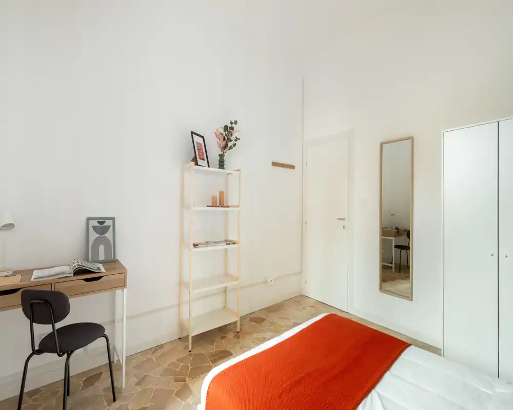 Camera da letto minimal e luminosa con un armadio bianco, scaffalatura semplice e letto con una calda coperta arancione. Linee pulite e luce naturale creano un’atmosfera moderna e accogliente, perfetta per affitti brevi.