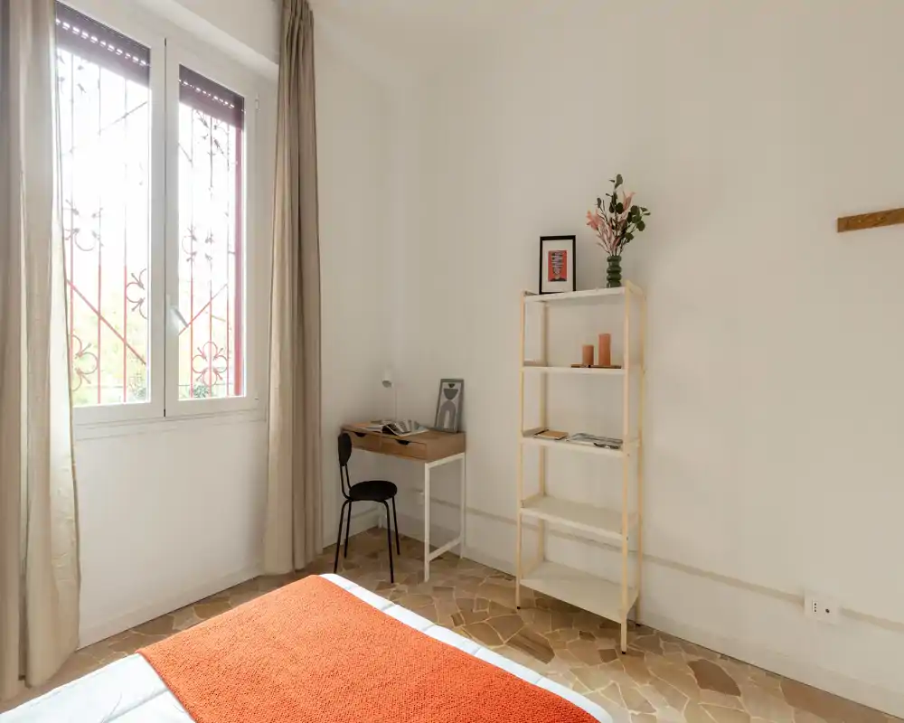 Camera da letto luminosa e minimal con letto ben rifatto, plaid arancione caldo e un piccolo angolo studio vicino alla finestra. Arredi semplici, mensole e tonalità neutre che creano un’atmosfera tranquilla.