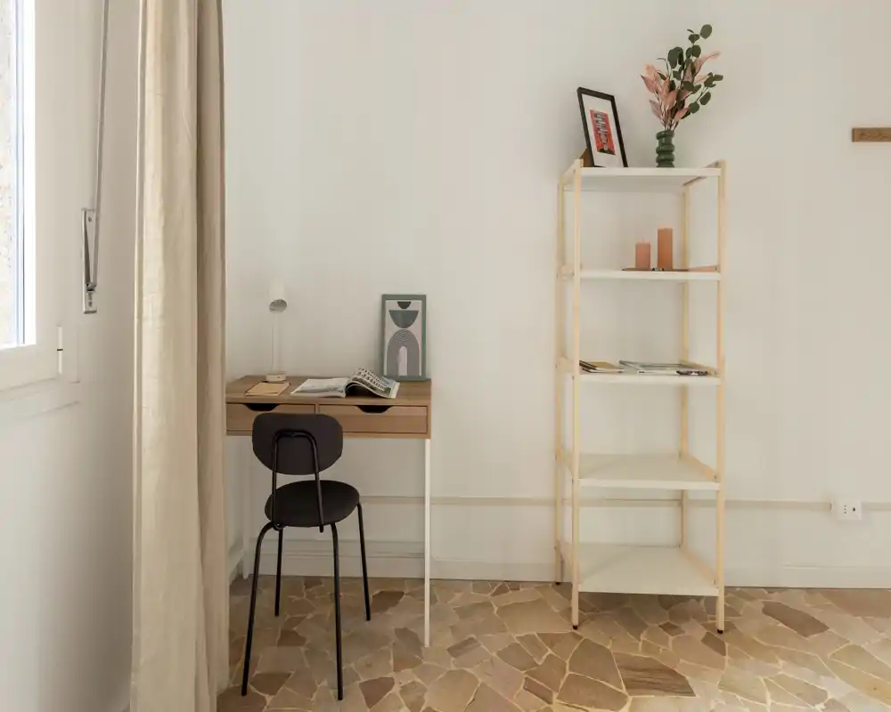 Spazio living minimal e luminoso con una piccola scrivania, sedia e scaffale aperto; tonalità neutre e luce naturale creano un angolo di lavoro tranquillo.