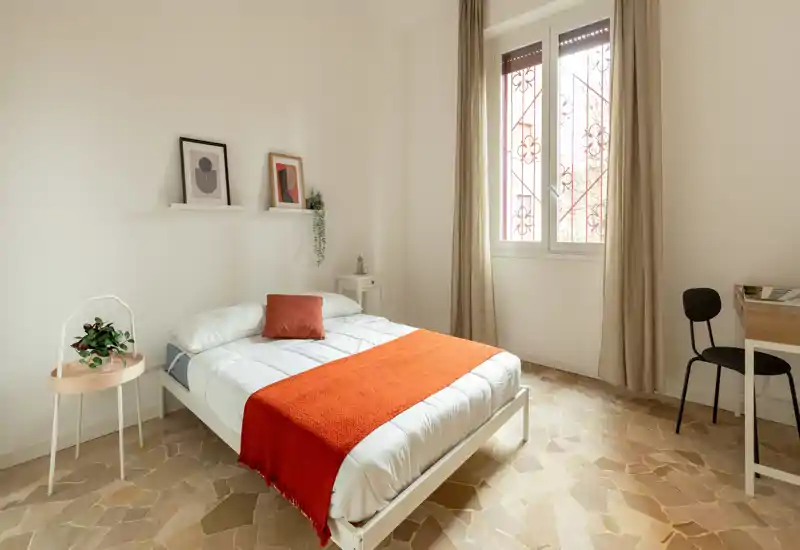 Camera da letto minimalista e luminosa con letto bianco, coperta arancione, comodino e una grande finestra che lascia entrare molta luce naturale.