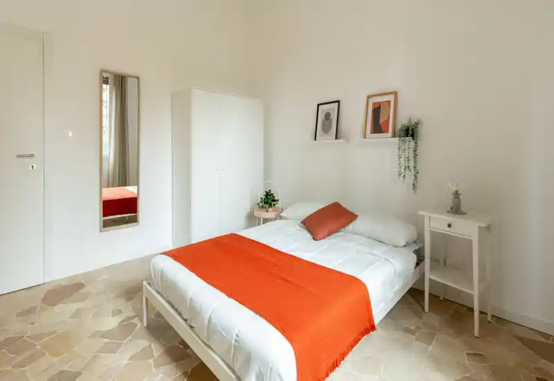 Camera da letto luminosa e pulita con letto matrimoniale in biancheria bianca e copriletto arancione. Arredamento minimale in stile scandinavo con piccolo armadio, comodino, quadri e specchio a figura intera che rendono l’ambiente accogliente.